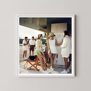 May include: A framed vintage photograph depicts a group of people at a tennis court. Several individuals are dressed in tennis attire, including white shorts and skirts. A woman in a white dress and a leopard print hat stands near a chart. Tennis rackets are visible.