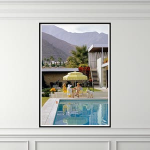 May include: A framed print of a mid-century modern scene. A group of people are gathered around a table under a yellow umbrella near a pool. Mountains and a modern house are in the background. A white dog is nearby.