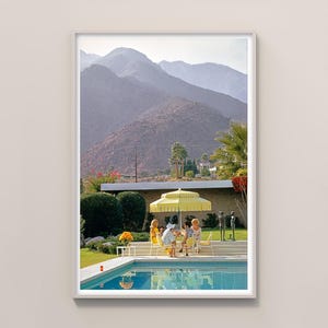 以下が含まれることがあります： レトロなプールサイドのシーンを特徴とする額入りのプリント。画像には、黄色いパラソルの下でテーブルに着席している人々のグループが写っており、プールと山の背景があります。このシーンは、ヴィンテージのミッドセンチュリーモダンな美学を連想させます。