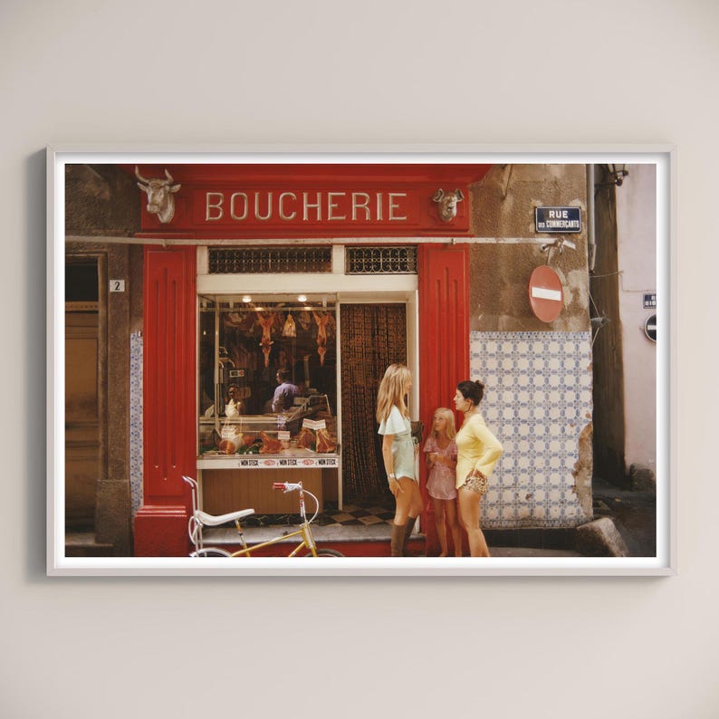 May include: A framed print of a French butcher shop, "Boucherie", with red trim and a yellow bicycle parked outside. Three people stand in front of the shop. The street sign reads "Rue des Commer&ccedil;ants".