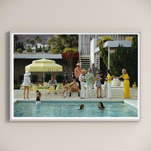 Könnte beinhalten: Gerahmter Druck eines Vintage-Fotos, das eine Poolparty zeigt. Menschen in stilvoller Kleidung versammeln sich um einen Swimmingpool, mit einem gelben Sonnenschirm und Palmen im Hintergrund. Das Bild erinnert an eine Freizeit-Szene aus der Mitte des Jahrhunderts.