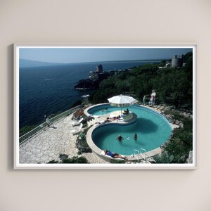 Puede incluir: Fotografía enmarcada de una piscina con un diseño único, con una isla central y una sombrilla blanca. La piscina está rodeada por un patio de piedra y tiene vistas al mar. Se ve a gente nadando y tomando el sol.