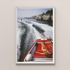 Puede incluir: Impresión enmarcada de una escena vintage con una lancha motora roja y blanca en el agua. Una persona en bikini rojo tomando el sol en el barco. Un esquiador acuático es visible a lo lejos, con una costa rocosa al fondo.