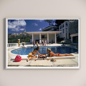 Könnte beinhalten: Ein gerahmter Druck eines Vintage-Fotos, das eine Szene am Pool zeigt. Zwei Personen spielen ein Spiel auf einer Matte, während sich andere am Pool entspannen. Eine rote Tasche und ein Strohhut befinden sich in der Nähe. Ein großes weißes Haus und eine Stadt auf einem Hügel sind im Hintergrund.