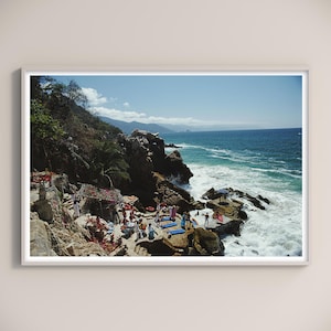 May include: A framed photograph of a coastal scene. The image captures a rocky shoreline with crashing waves under a bright blue sky. People are gathered on the rocks and in the water, enjoying the sunny day.