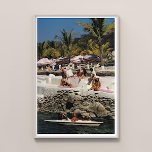 May include: A framed print depicting a vintage beach scene. Individuals are sunbathing on a white stone structure with pink mats, beneath striped umbrellas. A boat is on the water. Palm trees and a clear blue sky create a tropical atmosphere.