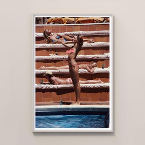 Pode incluir: Impressão emoldurada com uma fotografia retro de pessoas a tomar sol à beira da piscina. A imagem mostra pessoas em trajes de banho, relaxando em plataformas em camadas. A cena é banhada pela luz quente do sol, evocando o verão.