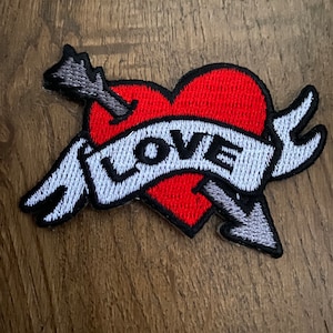 May include: Embroidered patch with a red heart pierced by a grey arrow. A white banner across the heart reads "LOVE" in black letters. The patch has a black border and is likely for clothing or accessories.