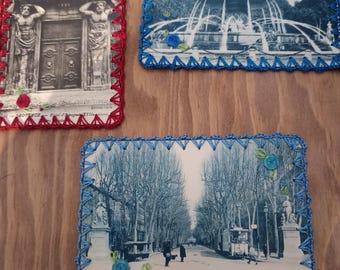 May include: Three vintage postcards with black and white images of European landmarks. Each postcard has a crocheted border in red or blue, with floral embellishments. The postcards are displayed on a wooden surface.