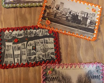 May include: Four rectangular vintage postcards featuring black and white photographs of Avignon, France. Each postcard is bordered with hand-crocheted yarn in green, orange, red, or pink, and decorated with small, stitched fabric flower accents.