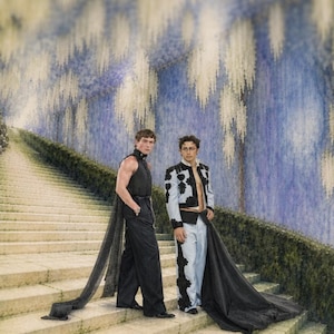 May include: Two people in stylish outfits stand on a stone staircase. One wears a black sleeveless top and pants with a long, flowing cape. The other is in a light blue jacket with black accents and a black cape.
