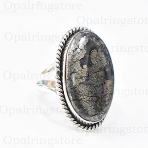 Puede incluir: Un anillo de plata con una piedra grande de forma ovalada. La piedra es de color gris oscuro con un acabado metálico brillante. El anillo tiene un diseño de cuerda alrededor del borde de la piedra.