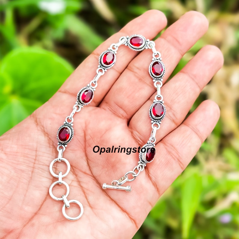 May include: A silver bracelet with red oval-shaped gemstones. The bracelet has a toggle clasp and a chain with a decorative design.