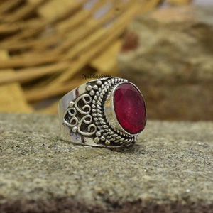 Könnte beinhalten: Ein silberner Ring mit einem verzierten Design und einem großen, ovalen Rubin-Edelstein. Der Ring liegt auf einer strukturierten, grauen Oberfläche.