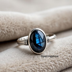 Op de afbeelding: Een zilveren ring met een ovale, donkerblauwe edelsteen met iriserende flitsen. De ring heeft een eenvoudige band met decoratieve details bij de steen. De tekst "Opalringstore" is zichtbaar.
