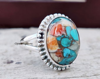 May include: A silver ring with a large oval-shaped gemstone. The gemstone is a mix of turquoise, orange, and white stones. The ring has a beaded edge.