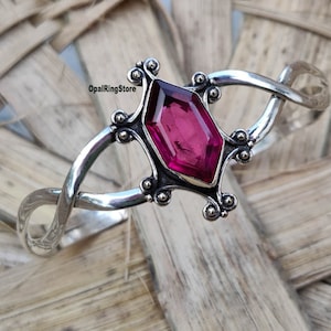 May include: A silver bangle bracelet with a pink gemstone set in a geometric design. The bracelet is twisted and has a delicate, ornate look.
