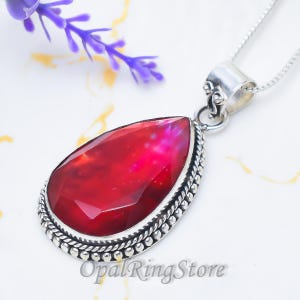 Puede incluir: Un collar con colgante en forma de lágrima que presenta una piedra preciosa de color rojo intenso facetada, engastada en un marco de plata esterlina pulida con un borde de cuentas. El colgante cuelga de una delicada cadena de plata.
