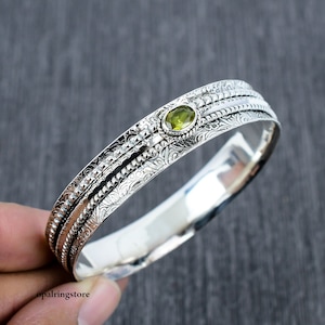 Op de afbeelding: Zilveren armband met een ingewikkeld bloemmotief en een groene edelsteen in het midden.