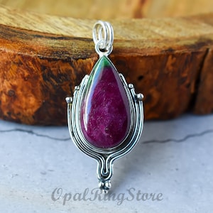 Peut inclure: Un pendentif en forme de larme avec une pierre précieuse verte et violette sertie dans un motif en filigrane argenté. Le pendentif est posé sur une surface en bois.
