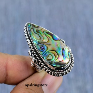 May include: A silver ring featuring a teardrop-shaped abalone shell inlay. The shell displays iridescent green, blue, and purple hues. The ring band is intricately crafted with a rope-like design.
