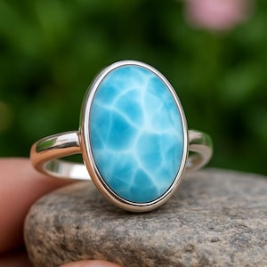 Könnte beinhalten: Ein Silberring mit einem ovalen Larimar-Edelstein. Der hellblaue Stein hat weiße Adern und ist in einem polierten Silberband gefasst. Der Ring wird auf einem grauen Stein präsentiert. Der Edelstein ist ca. 2,5 cm lang.