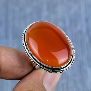 Könnte beinhalten: Ein silberner Ring mit einem ovalen Karneol-Edelstein. Der Ring hat ein detailliertes Silberband mit einem strukturierten Rand.