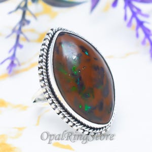 May include: A silver ring with a large, oval-shaped gemstone. The gemstone is primarily brown with flecks of green and blue. The ring has a detailed, textured border. The text "OpalRingStore" is visible at the bottom.