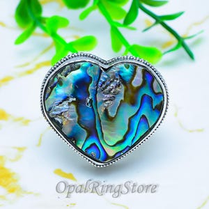 May include: A heart-shaped ring featuring an iridescent abalone shell inlay with swirling patterns of blue, green, and purple hues, set within a polished silver-toned metal frame with a beaded edge detail and the text OpalRingStore.