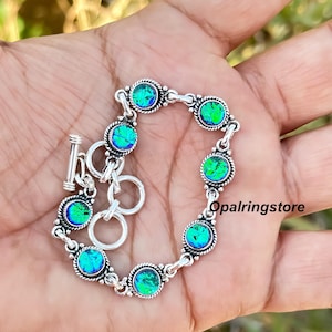 May include: Silver bracelet featuring round, iridescent blue-green stones. The bracelet has a toggle clasp and is composed of interconnected links. The stones are set in a decorative, beaded frame. The bracelet is displayed on a hand.