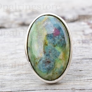 Könnte beinhalten: Ovaler Ring mit einem grün-blau-weißen Edelstein, der in einer silbernen Fassung gefasst ist. Der Edelstein hat ein wirbelndes Muster und einen Hauch von Rosa.