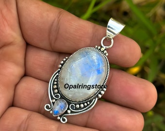 Op de afbeelding: Een zilveren hanger met een grote ovale maansteen en een kleinere maansteen accent. De hanger heeft een gedetailleerd ontwerp met kleine kralen en swirls. De tekst "Opalringstore" is zichtbaar in de afbeelding.