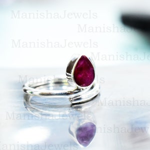 Può includere: Un anello in argento con una pietra preziosa rosso scuro a forma di goccia. L'anello ha un design unico a spirale con la pietra preziosa incastonata in una lunetta. L'anello è esposto su una superficie riflettente.