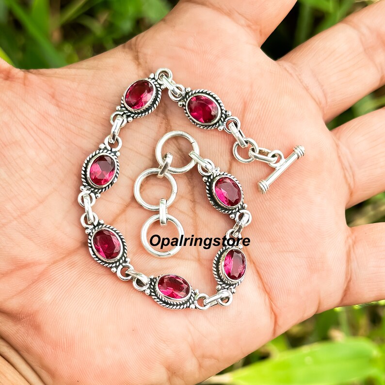 May include: A silver bracelet with a toggle clasp features seven oval-shaped pink gemstones set in silver bezels. The bracelet is on a hand.