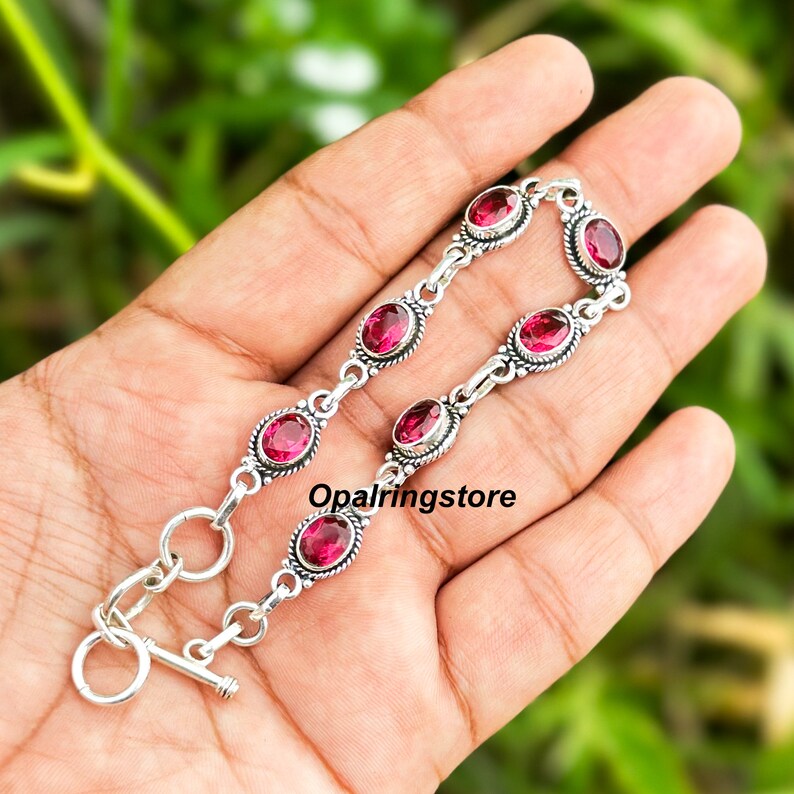 May include: A silver bracelet with a toggle clasp features a chain with oval-shaped pink gemstones set in silver.