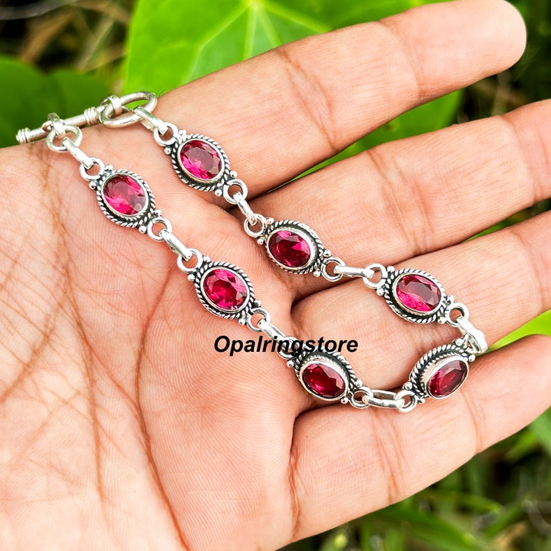 May include: A silver bracelet with a toggle clasp and oval-shaped pink gemstones. The bracelet is on a hand.