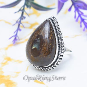 May include: A teardrop-shaped boulder opal ring set in a sterling silver band. The dark brown stone features iridescent green and gold flecks. The silver setting is detailed with a twisted rope border and a beaded edge surrounding the gemstone.