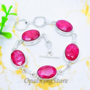 Può includere: Un bracciale in argento con pietre preziose rosse ovali e vibranti. Il bracciale ha una chiusura a toggle ed è composto da più maglie di pietre preziose. Le pietre sono incastonate in una montatura in metallo argentato. Il logo OpalRingStore è visibile.