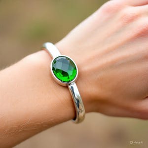 May include: A silver bracelet with a large, oval-cut, emerald-green gemstone. The gemstone is set in a polished metal bezel, and the bracelet has a simple, curved design. The bracelet is shown on a wrist.