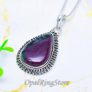 Puede incluir: Un colgante de plata con una cadena. El colgante en forma de lágrima presenta una piedra preciosa púrpura y verde, engastada en un marco plateado decorativo. El texto "OpalRingStore" es visible en la parte inferior.
