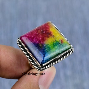 May include: A square-shaped ring with a colourful gemstone set in a silver band. The gemstone has a rainbow of colours, including red, orange, yellow, green, blue, and purple. The band has a rope-like design.