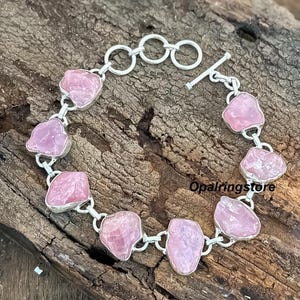 Op de afbeelding: Een zilveren armband met onregelmatige, lichtroze edelstenen. De stenen zijn gezet in zilver en verbonden door zilveren schakels. De armband heeft een tuimelsluiting. De tekst "Opalringstore" is zichtbaar.