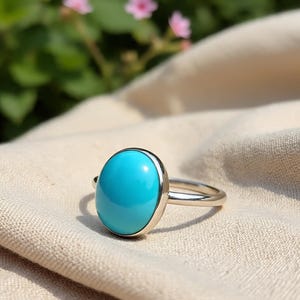May include: A silver ring featuring a large, oval-shaped, bright turquoise stone. The gemstone is set in a polished silver band, creating a striking contrast. The ring is displayed on a beige fabric, with soft focus greenery in the background.