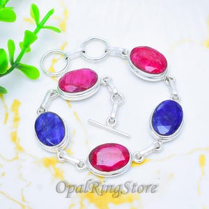 Può includere: Un bracciale in argento con pietre preziose ovali rosse e blu. Le pietre sono incastonate in castoni individuali e collegate da maglie d'argento. Ha una chiusura a barretta. Il testo "OpalRingStore" è visibile.
