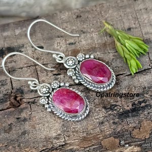 以下が含まれることがあります： オーバル型でファセットカットのルビーレッドの宝石が付いたシルバーのイヤリング。イヤリングには装飾的なシルバーの縁取りとフック留めが付いています。背景は、小さな緑色の小枝のある木製の表面です。