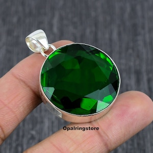 Puede incluir: Un colgante redondo de piedra preciosa verde con un engaste de plata. El colgante está facetado y tiene un alto pulido. El colgante está en un dedo.