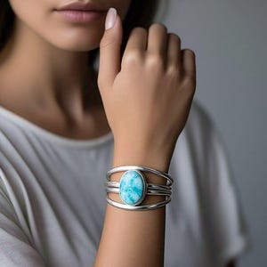 Könnte beinhalten: Ein silbernes Armreif-Armband mit einem großen, ovalen, hellblauen Edelstein. Das Armband hat ein mehrreihiges Design. Der Edelstein hat ein marmoriertes Aussehen mit Blau- und Weißtönen.