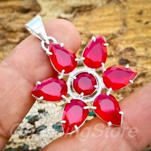 Pode incluir: Pingente de prata com design floral, cravejado com sete pedras preciosas vermelhas. Seis pedras têm formato de gota e uma é redonda no centro. O pingente tem uma moldura de metal prateado e uma argola na parte superior para uma corrente.