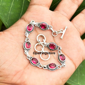 May include: A silver bracelet with a toggle clasp and pink oval-shaped gemstones. The bracelet is on a person's hand.