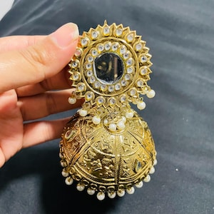 May include: Gold-coloured earring with a sunburst design, a central mirror, and small white pearls. The earring has intricate embossed patterns. It is held against a dark background.